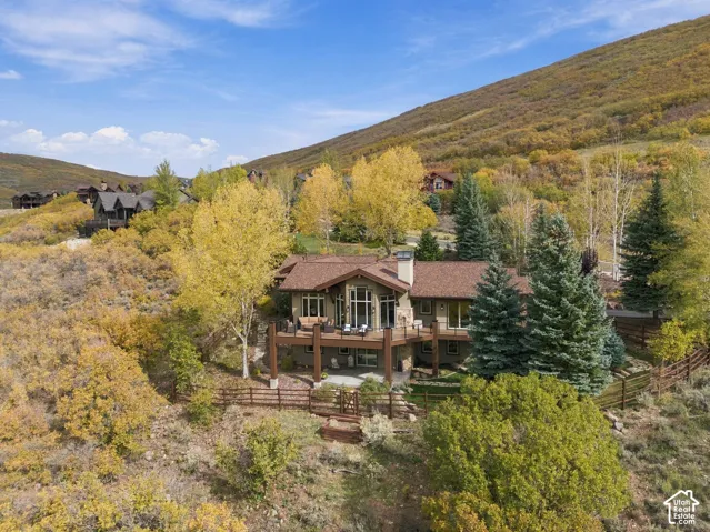 Multi-level residence featuring a wooden deck, stone-accented chimney, expansive windows, tiered patio, and perimeter wood fencing, situated against a mountain backdrop