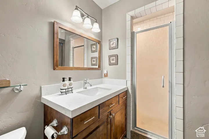 Wood-finish cabinetry vanity with white stone countertop, undermount sink, three-light wall-mounted fixture, and shower enclosure with tile surround