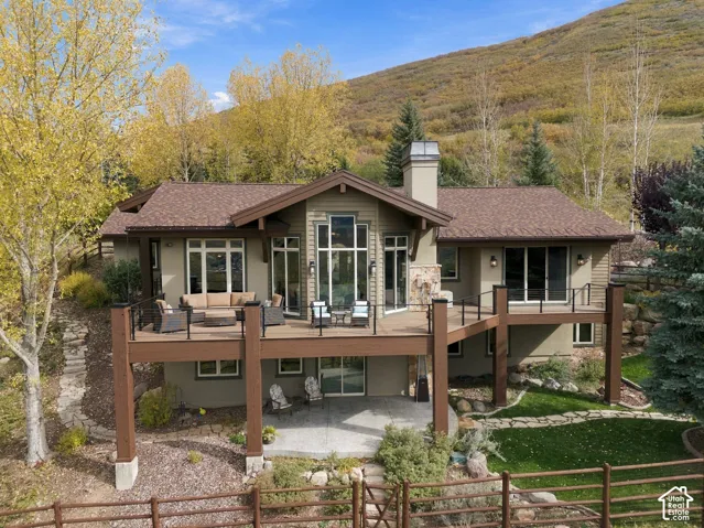 Multi-level exterior featuring wood-finish siding, a raised deck with metal railings, a stone fireplace chimney, and a concrete patio set against a mountain backdrop