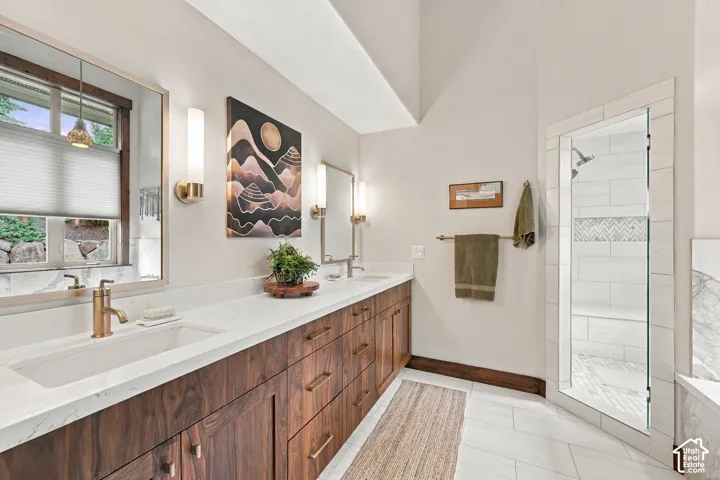 Dual-vanity cabinetry with wood-grain finish, white stone-surface countertops, brass-toned fixtures, rectangular wall-mounted lighting, and a glass-enclosed shower featuring tiled inlay and bench seating
