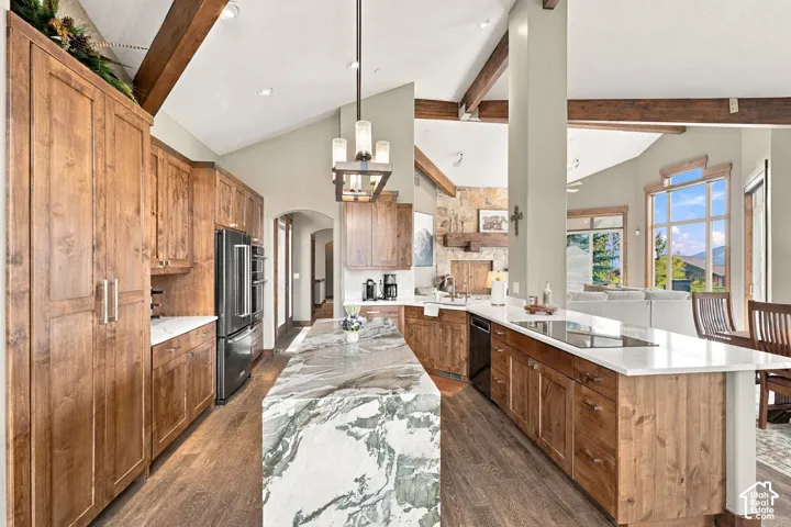 Kitchen featuring custom wood cabinetry, stone-topped waterfall island, vaulted ceilings with exposed wood beams, and built-in stainless steel appliances