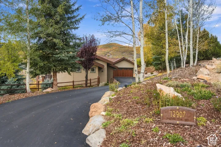 Asphalt driveway leading to a multi-car garage, split-rail fencing, mature evergreen and aspen trees, and rock-scaped terrain