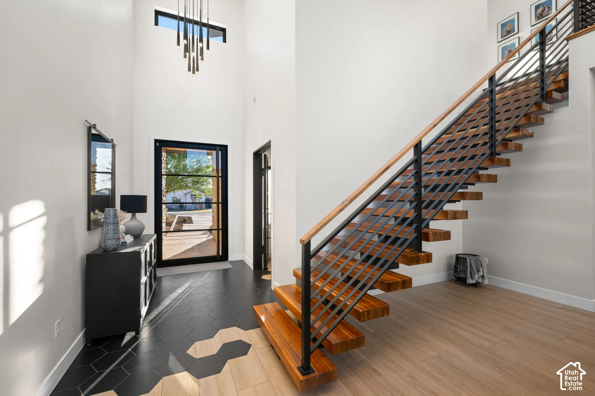 Foyer featuring high ceilings, black hexagonal floor tile transitioning to wood-finish flooring, a floating staircase with metal railings, and a multi-light pendant fixture