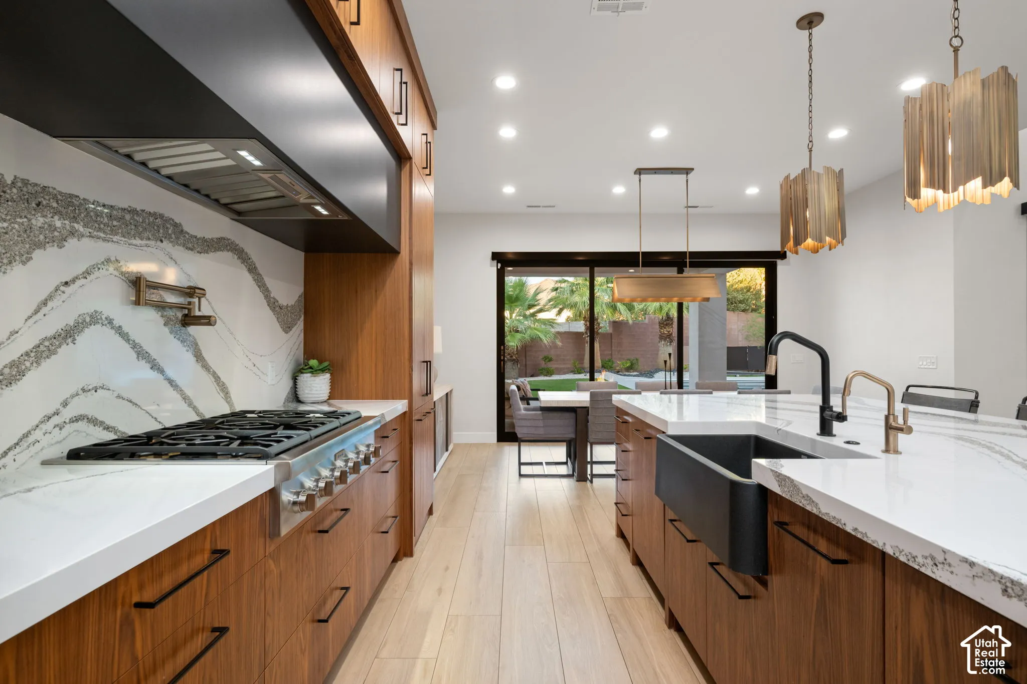 Kitchen featuring wood-finish cabinetry, white slab countertops with veining, a stainless steel gas cooktop with matching range hood, a matte black farmhouse sink with mixed-metal faucets, and wood-finish flooring