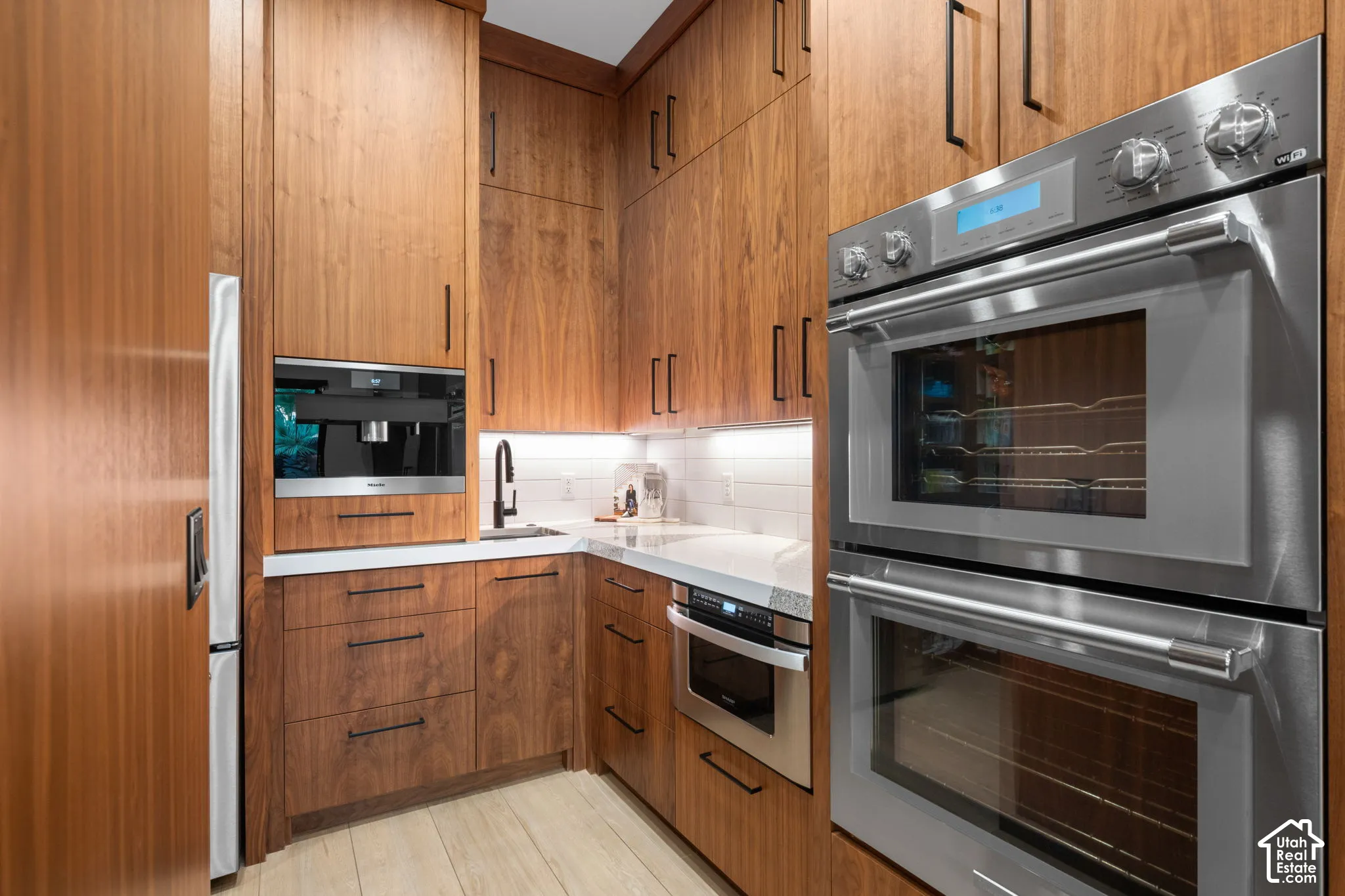 Custom wood-finish cabinetry with matte black hardware, stainless steel double wall ovens, integrated coffee system, under-cabinet lighting, and stone countertops