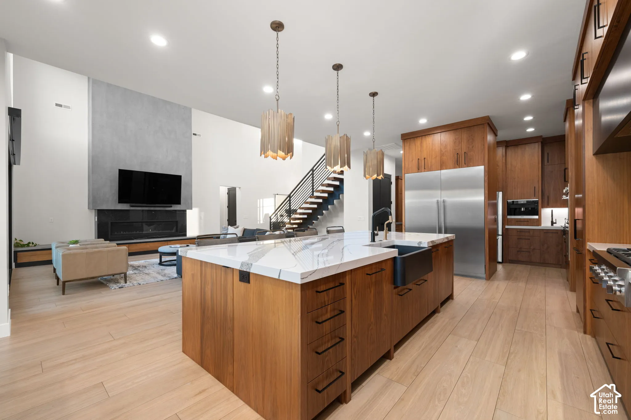 Kitchen featuring wood cabinetry, a large waterfall-edge island with a farmhouse sink, stainless steel appliances, and wood-finish flooring, complemented by a floor-to-ceiling fireplace surround
