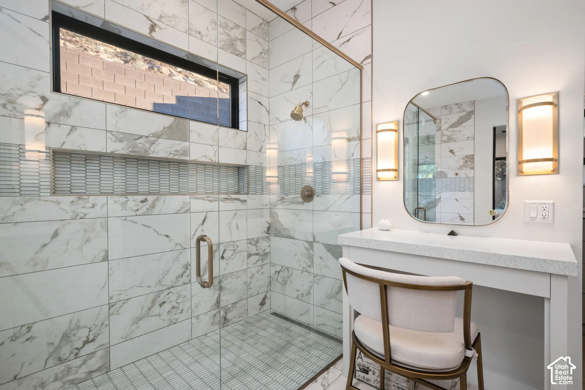 Walk-in shower featuring large-format stone-patterned tile, a recessed horizontal niche with decorative tile inlay, a glass enclosure, and a gold-finish rainfall showerhead, complemented by a built-in vanity with a stone countertop and gold-finish wall sc