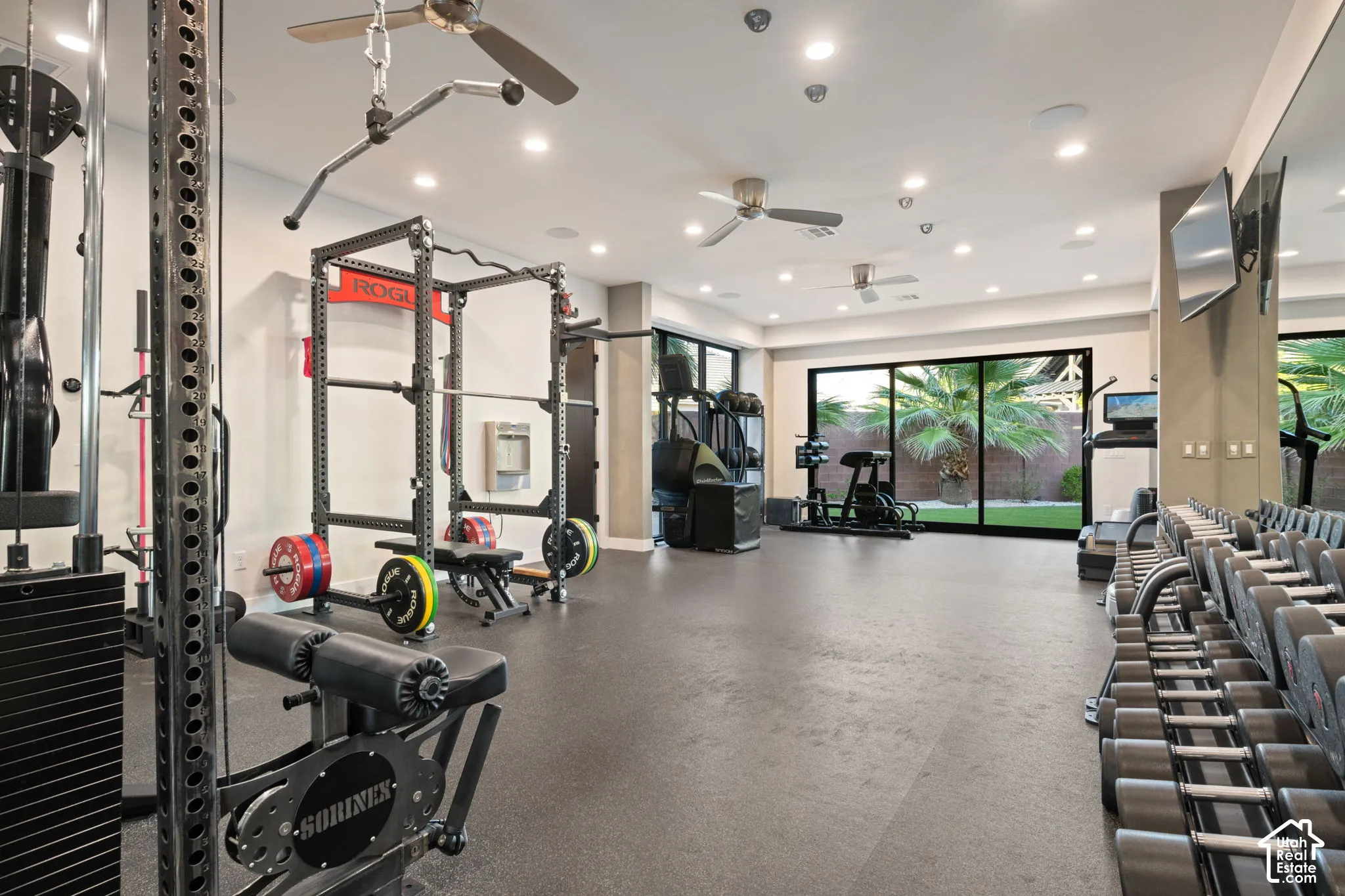 Spacious fitness room featuring rubberized flooring, recessed lighting, multi-blade ceiling fans, wall-to-wall mirror, and floor-to-ceiling sliding glass doors
