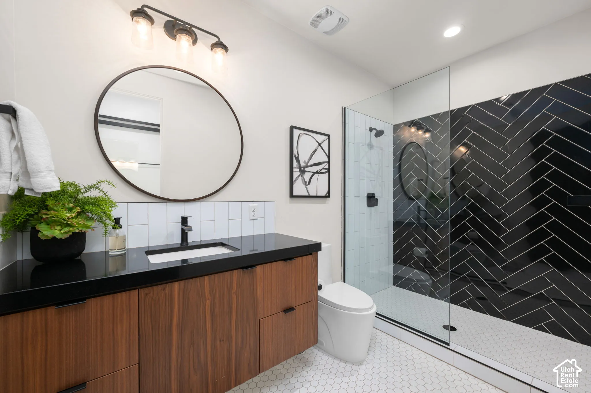 Wood-finish vanity with a dark countertop and integrated rectangular sink, round mirror with a thin dark frame, three-light wall fixture, and walk-in shower featuring a glass enclosure and black herringbone tile surround