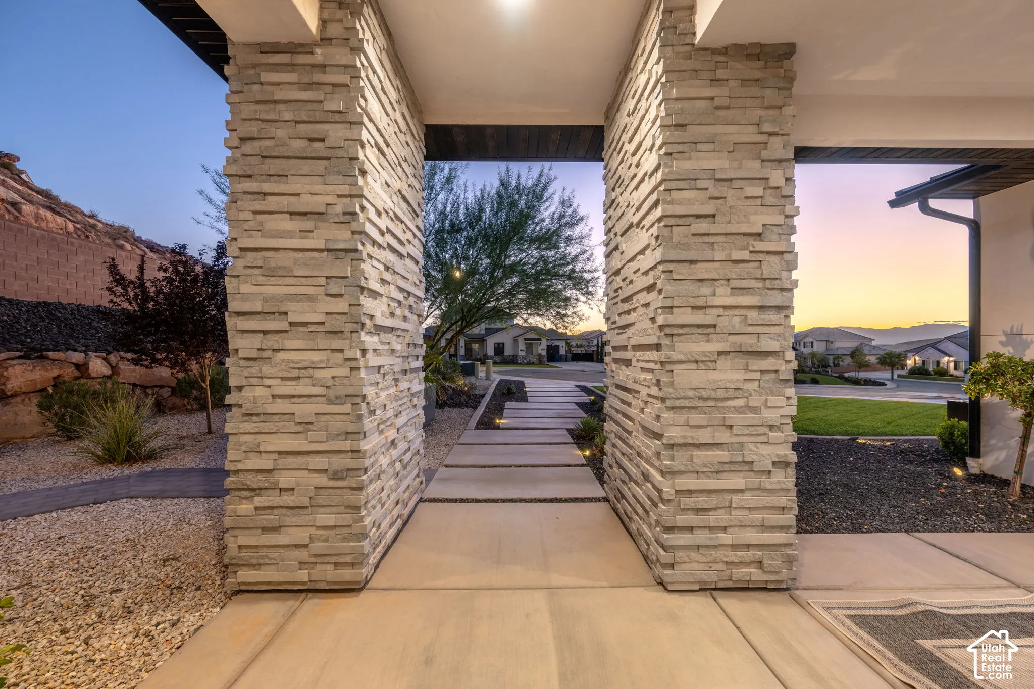 Covered entryway featuring dual stacked-stone columns, concrete slab walkway, and integrated landscape lighting