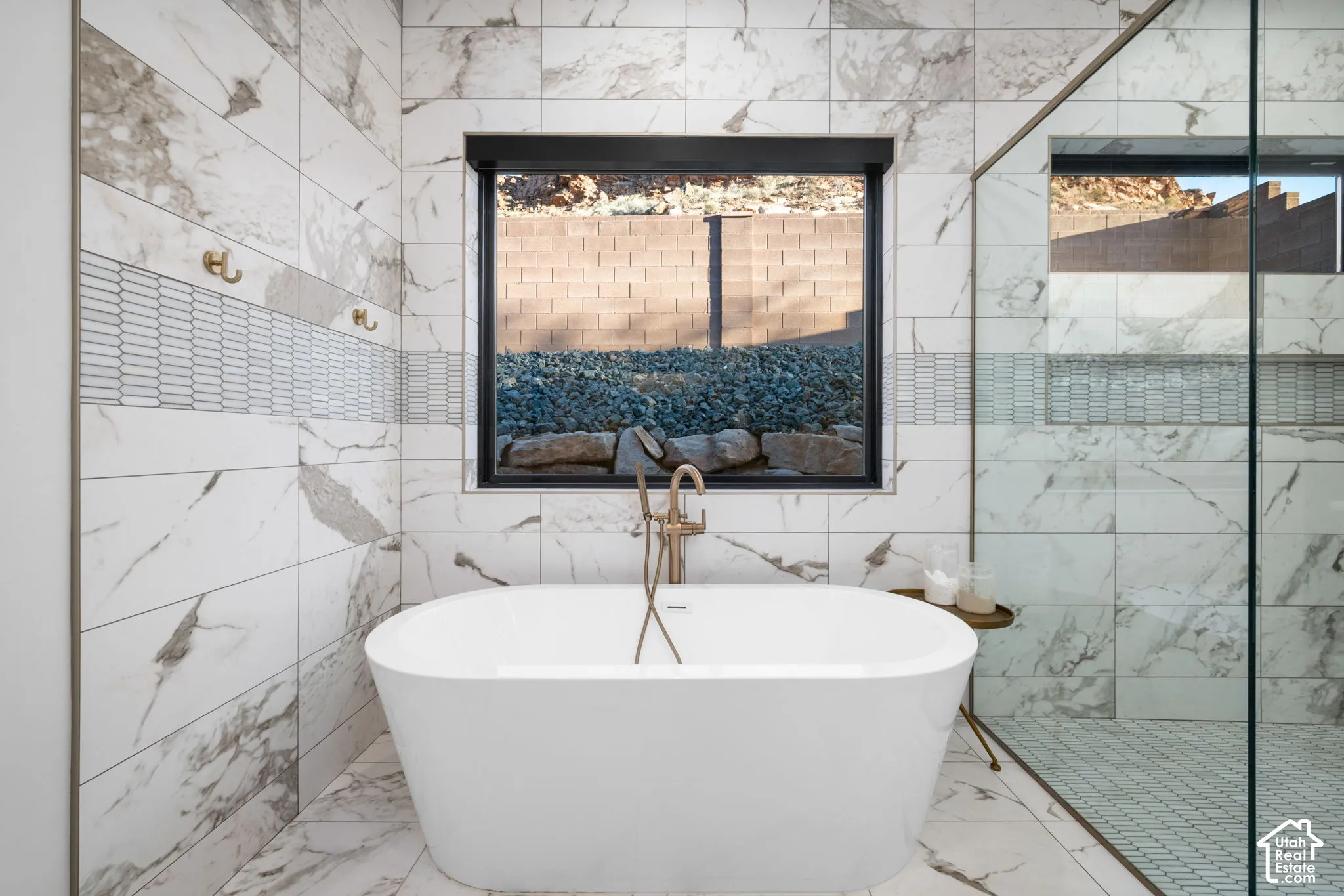 Freestanding soaking tub with a floor-mounted brass faucet, large-format stone-patterned tile wall cladding, glass-enclosed shower, mosaic tile accent border, and dual brass wall-mounted hooks
