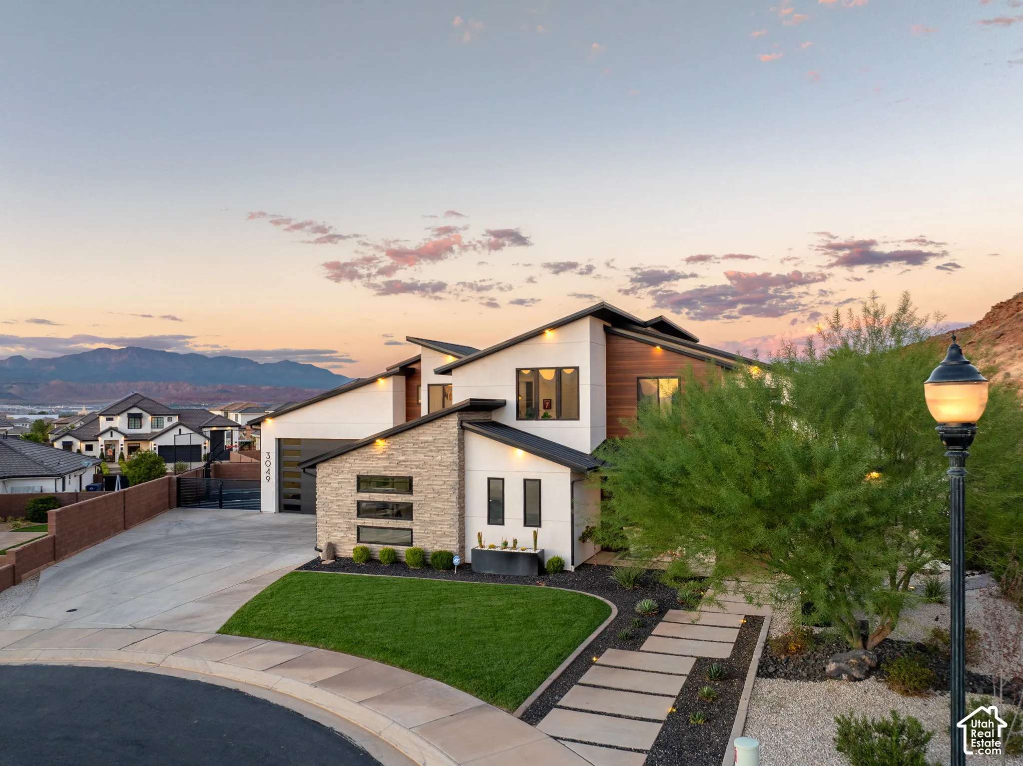 Contemporary architectural exterior featuring white smooth-finish siding, stacked stone accents, horizontal wood-finish cladding, a multi-pitch roofline, and a paved walkway with mature desert landscaping