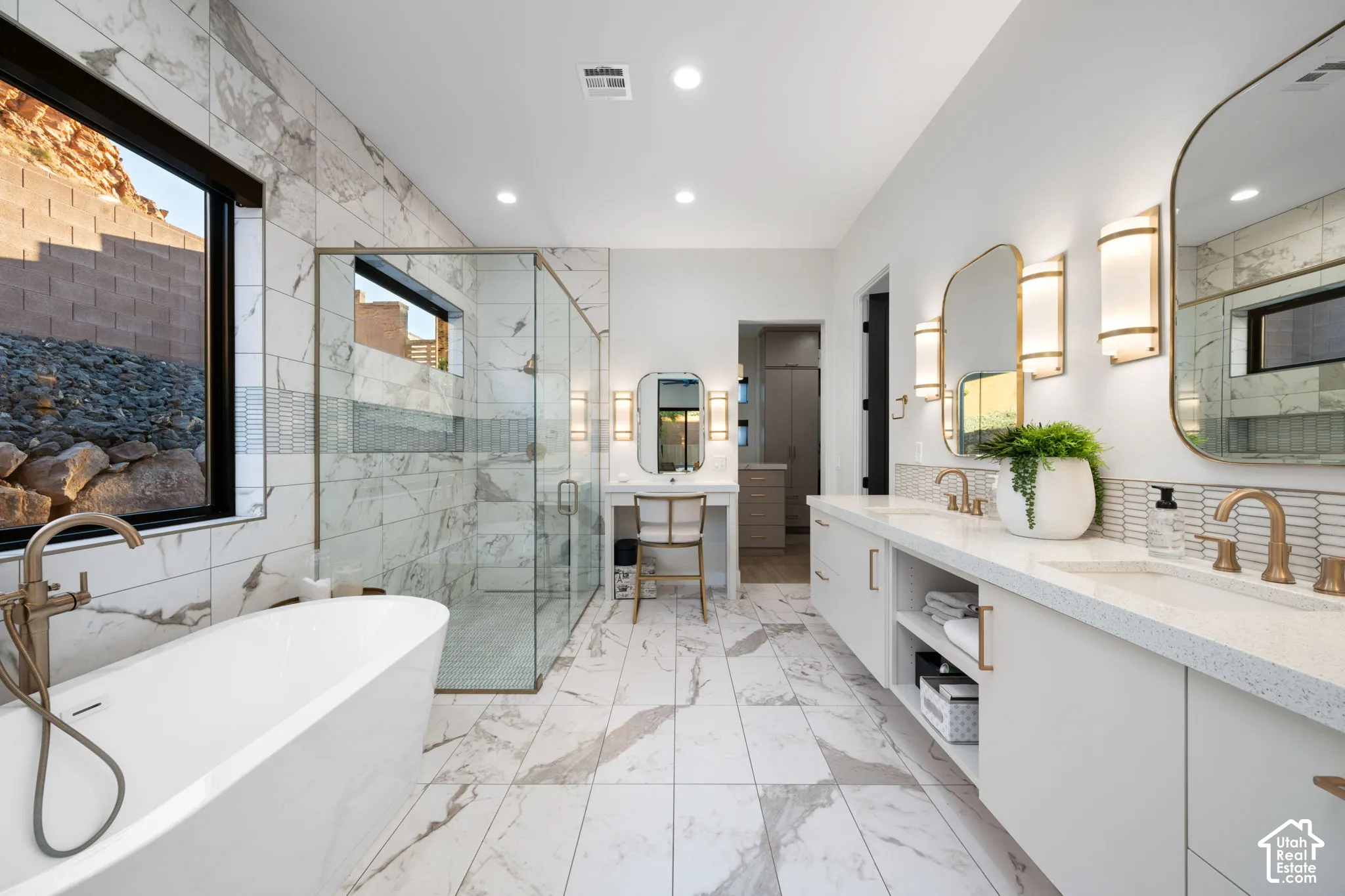 Freestanding soaking tub with brass fixtures, glass-enclosed walk-in shower, dual-sink vanity with stone-finish countertops, and large-format tile flooring