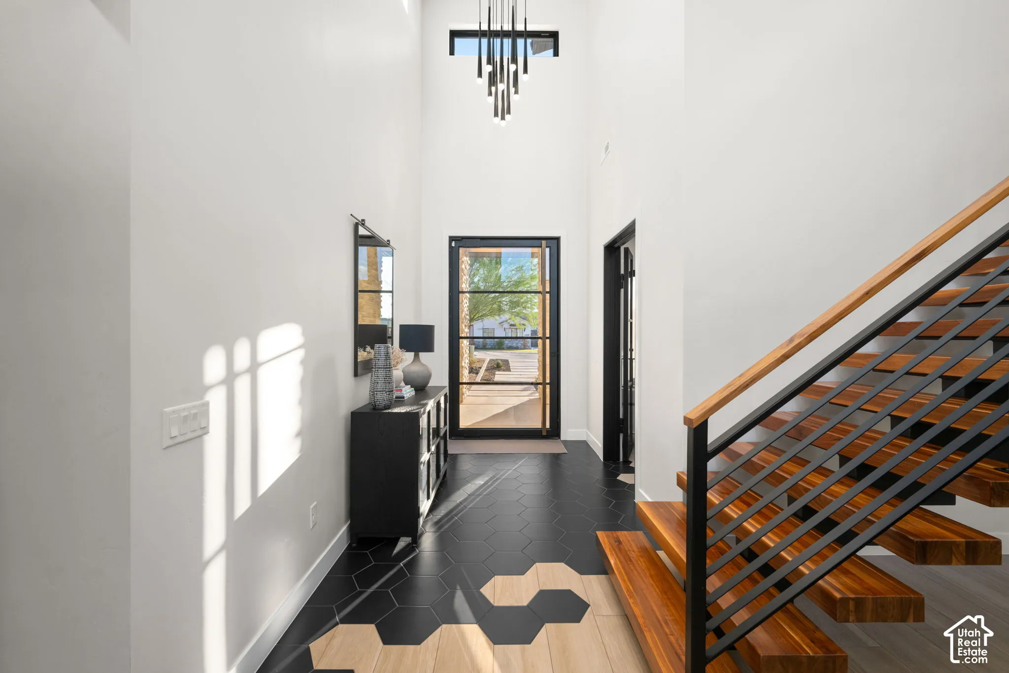 Double-height ceiling, open-riser staircase with wood-finish treads, black metal railing, patterned hexagonal tile flooring, and multi-pane glass entry door