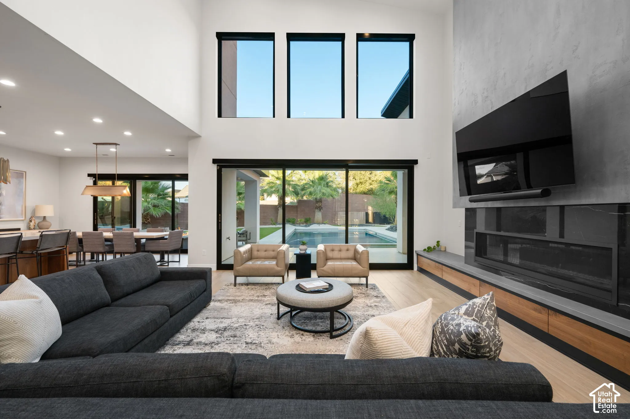 Double-height ceiling with expansive windows, floor-to-ceiling fireplace featuring concrete and wood-finish detailing, recessed lighting, and large glass sliding doors providing external exposure to the pool area