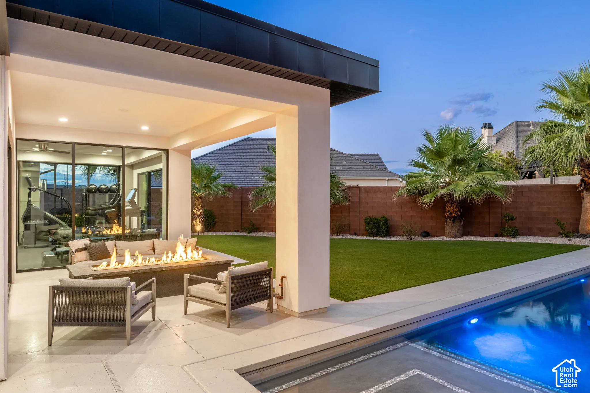 Covered patio featuring a built-in fire feature, in-ground pool with stone coping, synthetic turf lawn, and solid masonry perimeter wall