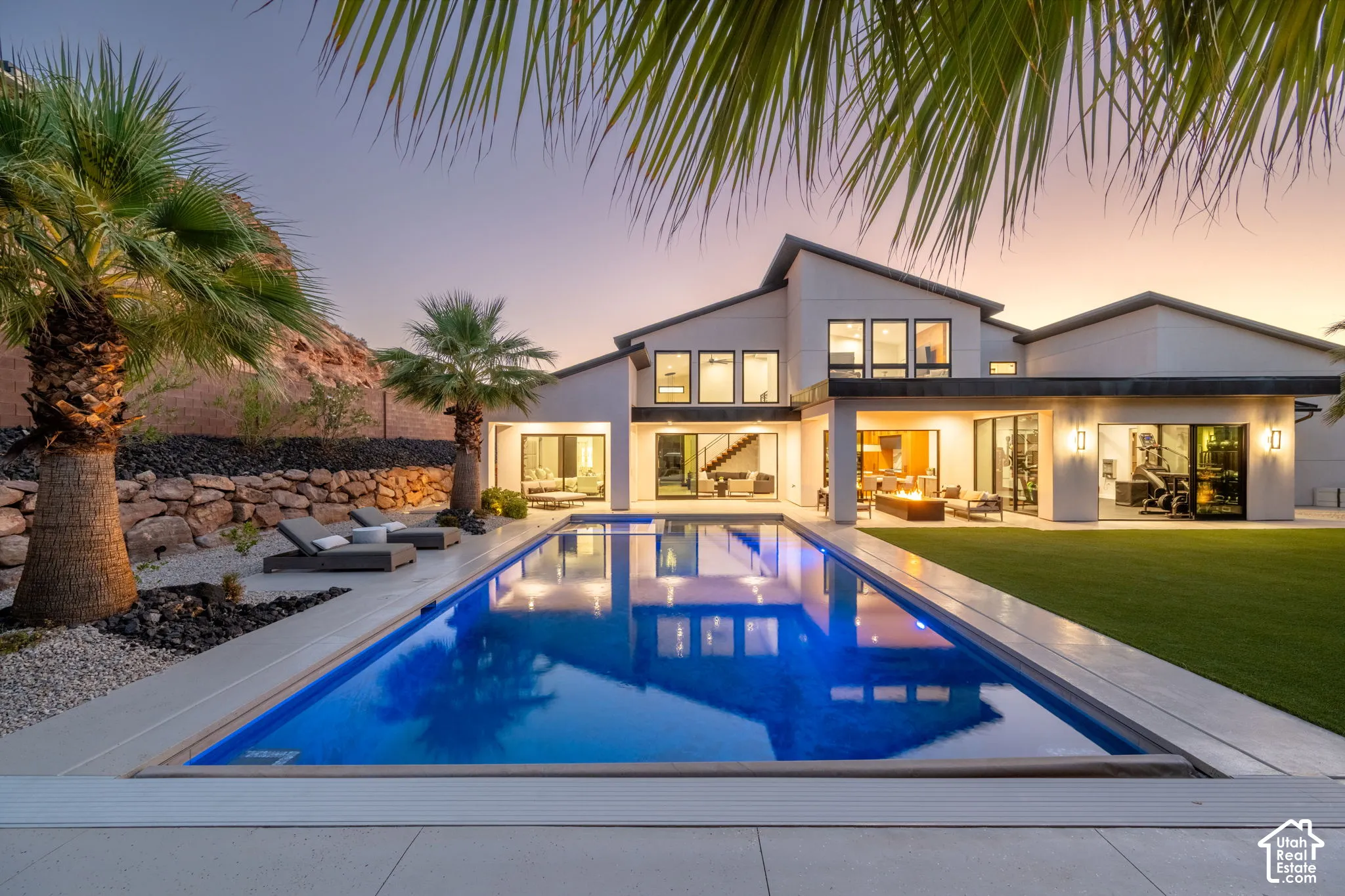 Contemporary rear exterior featuring a rectangular inground pool, manicured lawn, stacked stone retaining wall, and covered patio area with integrated lighting