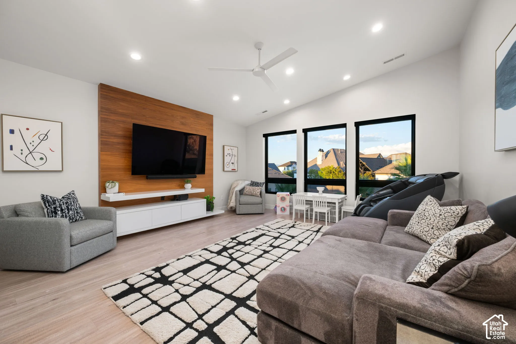 Living area featuring a vaulted ceiling, wood-finish flooring, recessed lighting, floor-to-ceiling wood-accent wall with a floating media console, and large windows with black frames
