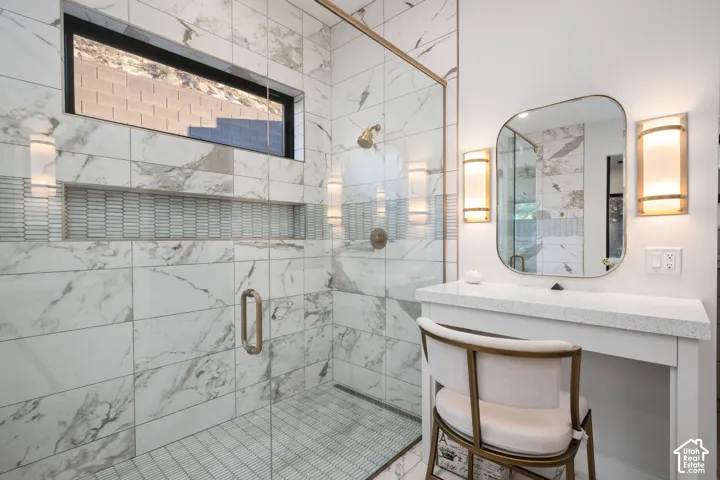 Walk-in shower featuring large-format stone-patterned tile, a recessed horizontal niche with decorative tile inlay, a glass enclosure, and a gold-finish rainfall showerhead, complemented by a built-in vanity with a stone countertop and gold-finish wall sc