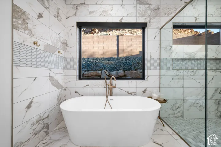 Freestanding soaking tub with a floor-mounted brass faucet, large-format stone-patterned tile wall cladding, glass-enclosed shower, mosaic tile accent border, and dual brass wall-mounted hooks
