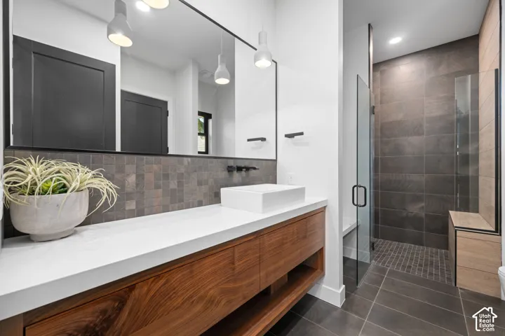 Floating wood-finish vanity with a rectangular vessel sink and solid-surface countertop, full-width mirror, mosaic tile backsplash, matte black wall-mounted faucet, and glass-enclosed shower with large-format tile walls