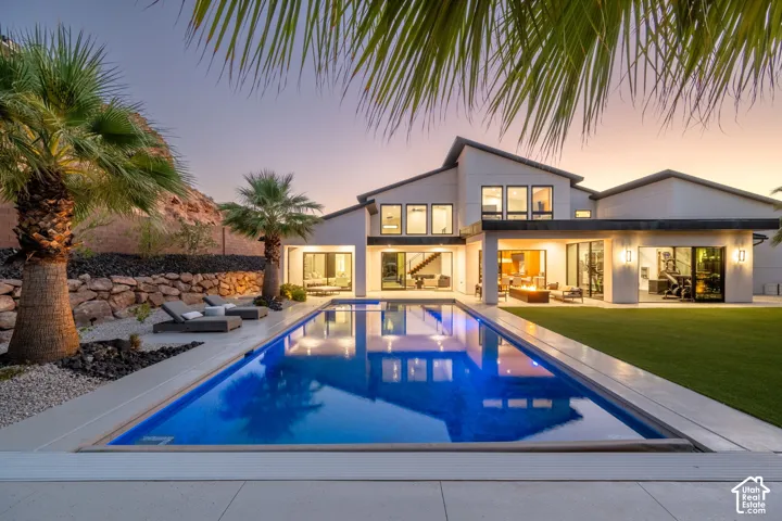 Contemporary rear exterior featuring a rectangular inground pool, manicured lawn, stacked stone retaining wall, and covered patio area with integrated lighting