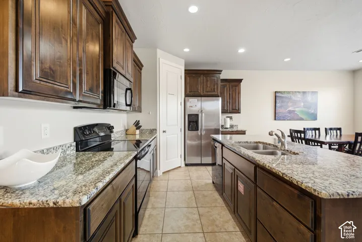 Kitchen featuring dark wood cabinetry, natural stone countertops, a center island with an undermount dual-basin sink, tile flooring, and stainless steel appliances