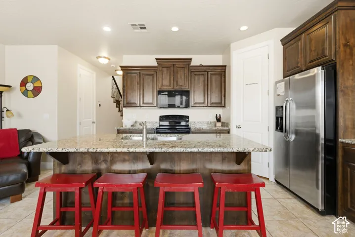 Kitchen featuring stained wood cabinetry, a central island with stone-finish countertops, stainless steel refrigerator, black range, and tile flooring