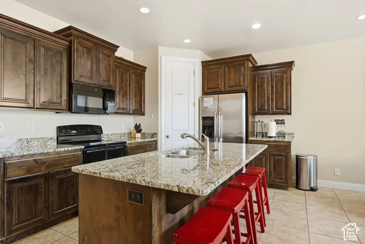 Kitchen featuring stained wood cabinetry, stone-surface countertops, a center island with an integrated dual-basin sink, stainless steel appliances, and tile flooring