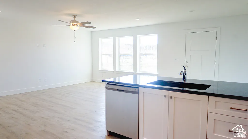 Kitchen island featuring a dark stone countertop, undermount sink, and stainless steel dishwasher, complemented by light wood-finish flooring, ceiling fan, and triple windows