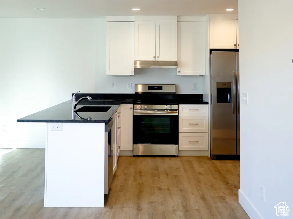 Kitchen featuring white shaker-style cabinetry, dark stone countertops, stainless steel appliances, a peninsula with an undermount sink, and wood-finish flooring