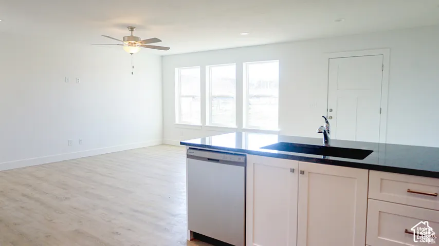 Kitchen island featuring a dark stone countertop, undermount sink, and stainless steel dishwasher, complemented by light wood-finish flooring, ceiling fan, and triple windows