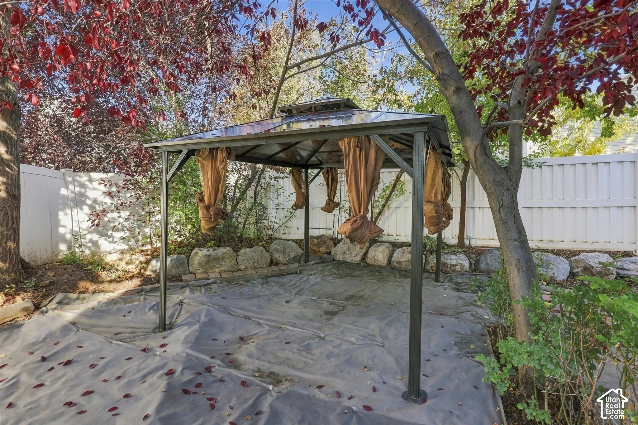 Backyard featuring a permanent metal-framed gazebo with a hard-top roof, bordered by natural stone accents and a white privacy fence, complemented by mature trees
