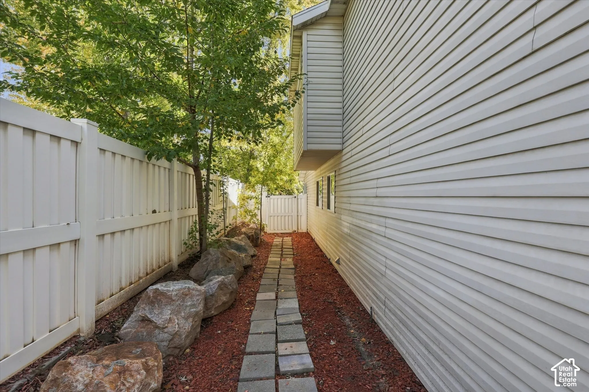 Side yard featuring horizontal vinyl siding, a white privacy fence, stone boulder accents, a paved stone walkway, and red mulch ground cover