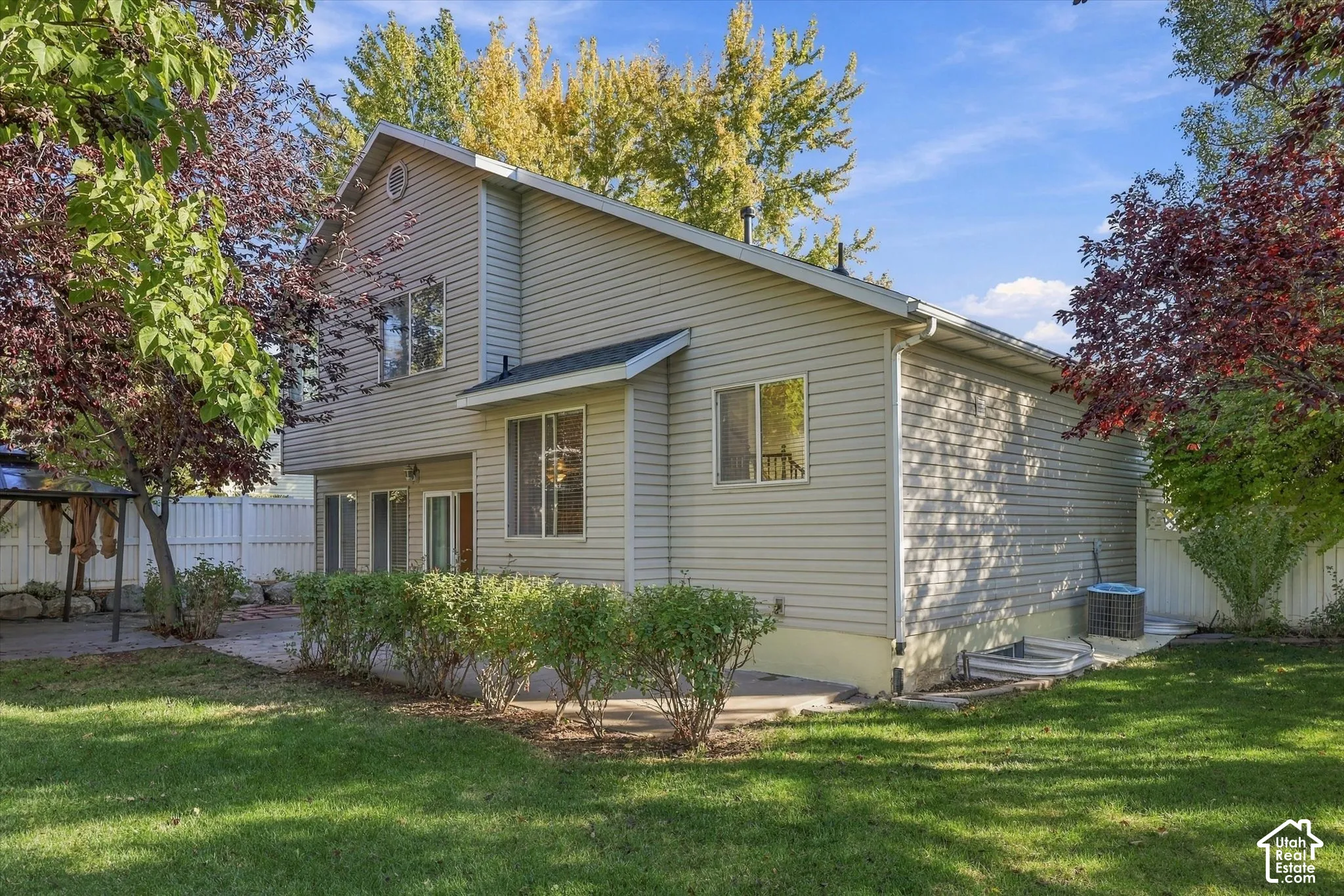 Two-story exterior featuring light-toned horizontal siding, a concrete patio, established perimeter landscaping, and a fenced yard