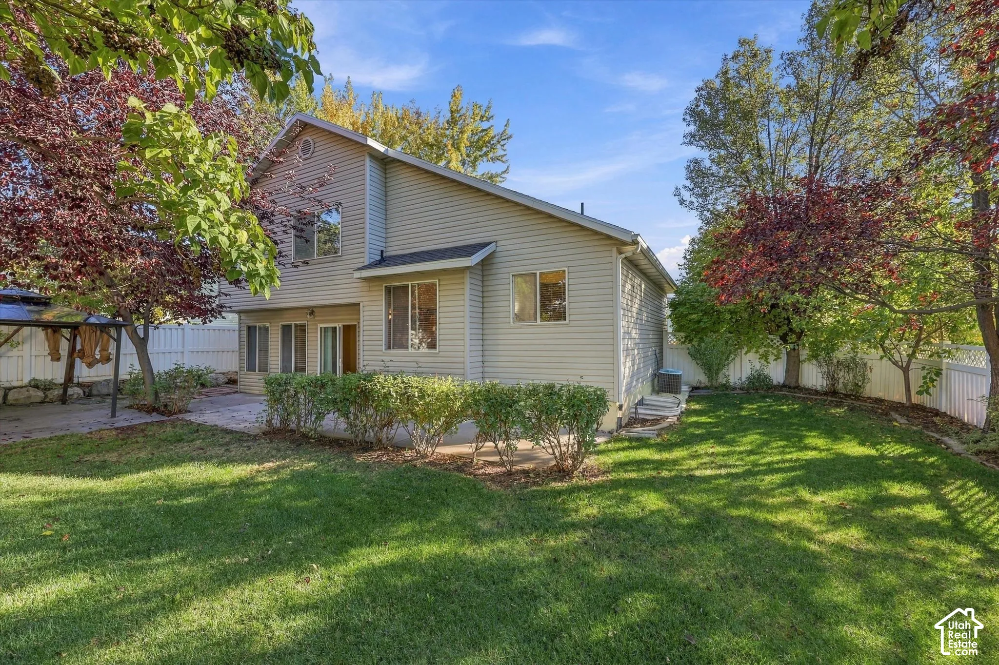 Two-story residence with neutral siding, paved patio, mature trees, and white vinyl privacy fencing