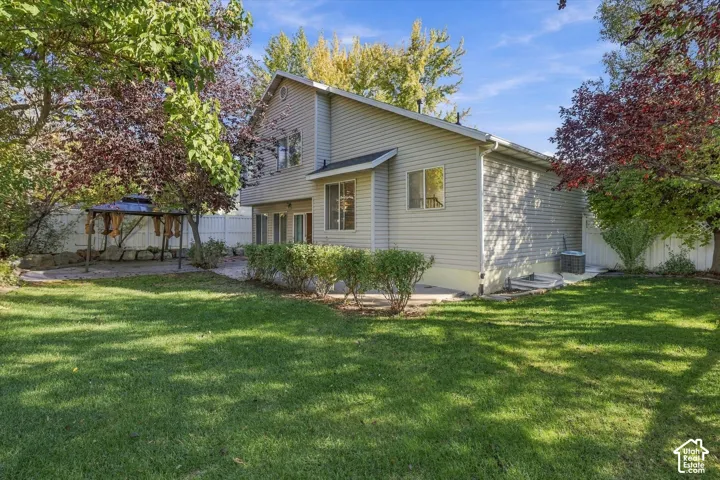 Two-story residence with light-colored siding, featuring a spacious grass lawn, mature trees, a concrete patio, and white perimeter fencing