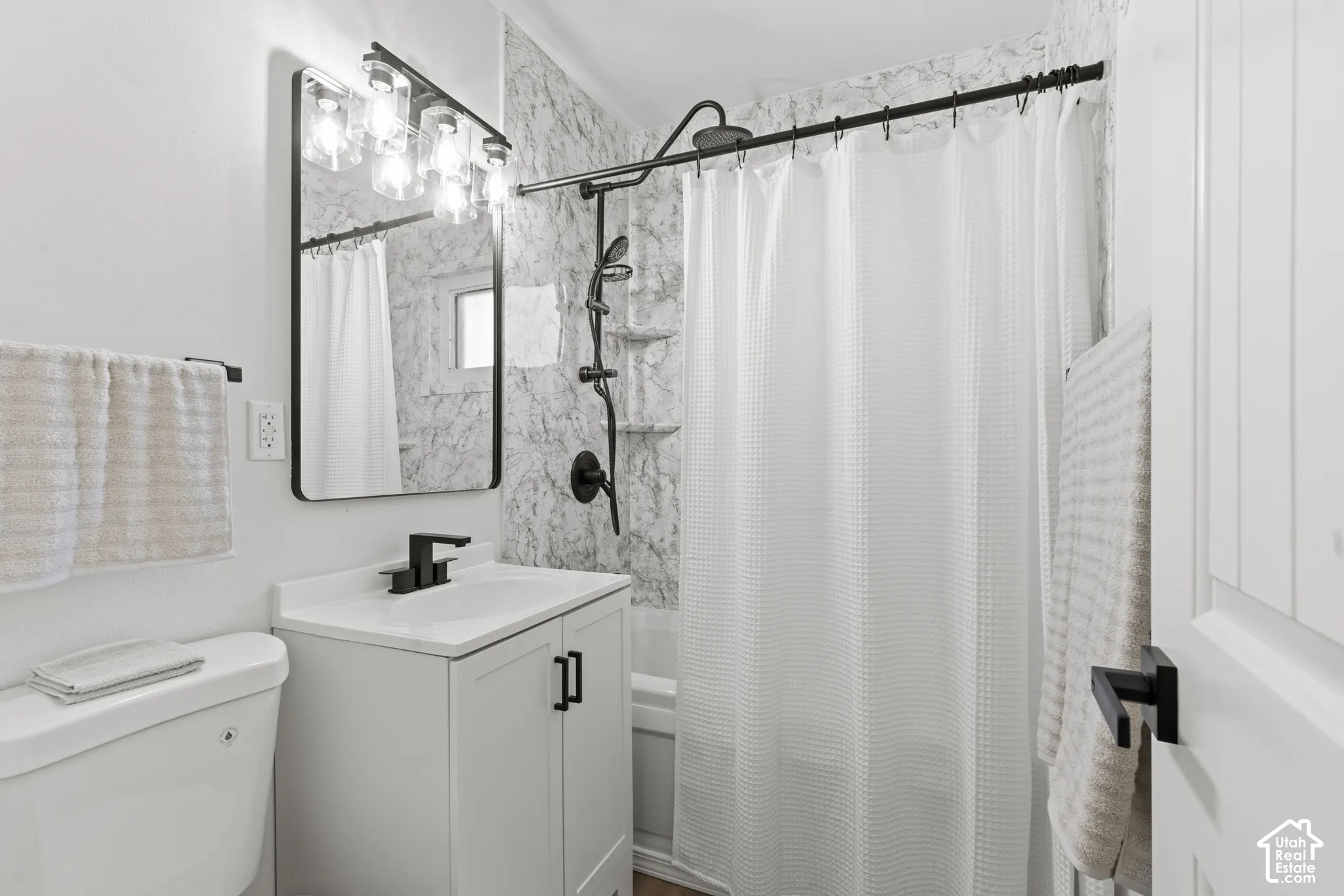 Bathroom featuring a vanity with a white countertop, wall-mounted rectangular mirror, matte black faucet and hardware, and shower surround with stone-patterned tile