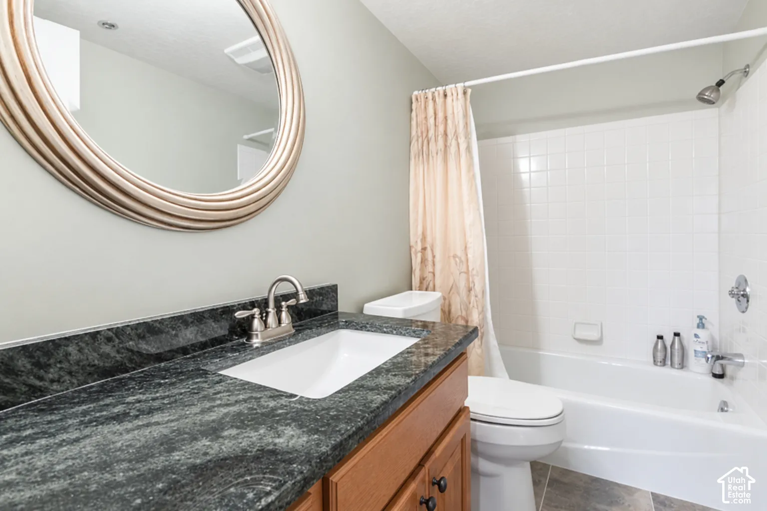 Bathroom featuring a wood-finish vanity with a dark stone-surface countertop, an oval wall-mounted mirror with a decorative frame, and a white porcelain tub-shower combination with tiled surround