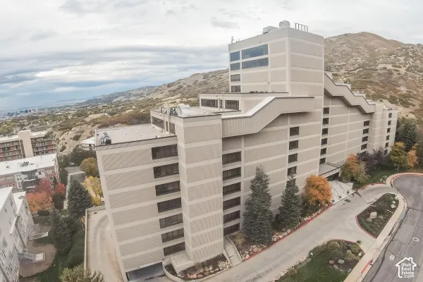 Multi-story building exterior featuring a stepped structural profile, concrete panel siding, mountain terrain, and an asphalt driveway with integrated landscaping