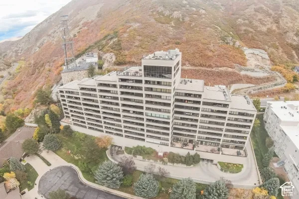 Multi-story residential building featuring terraced balconies, a rooftop deck, paved circular driveway, and mature coniferous landscaping, situated against a mountainous backdrop