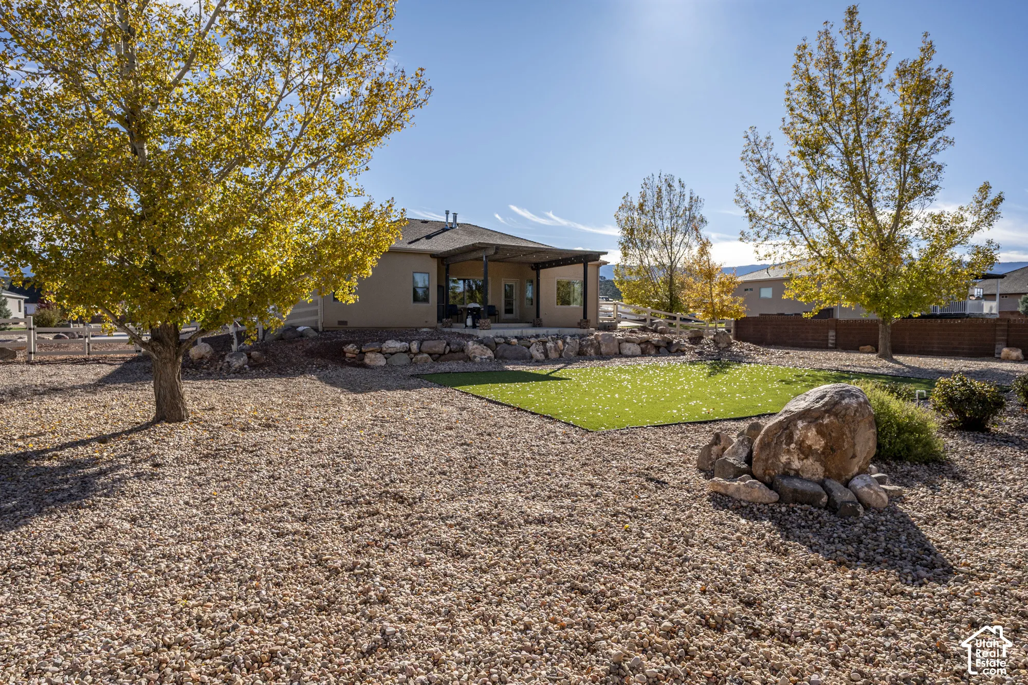 Backyard featuring a covered patio, tiered natural stone retaining wall, synthetic grass lawn, and decorative gravel landscaping