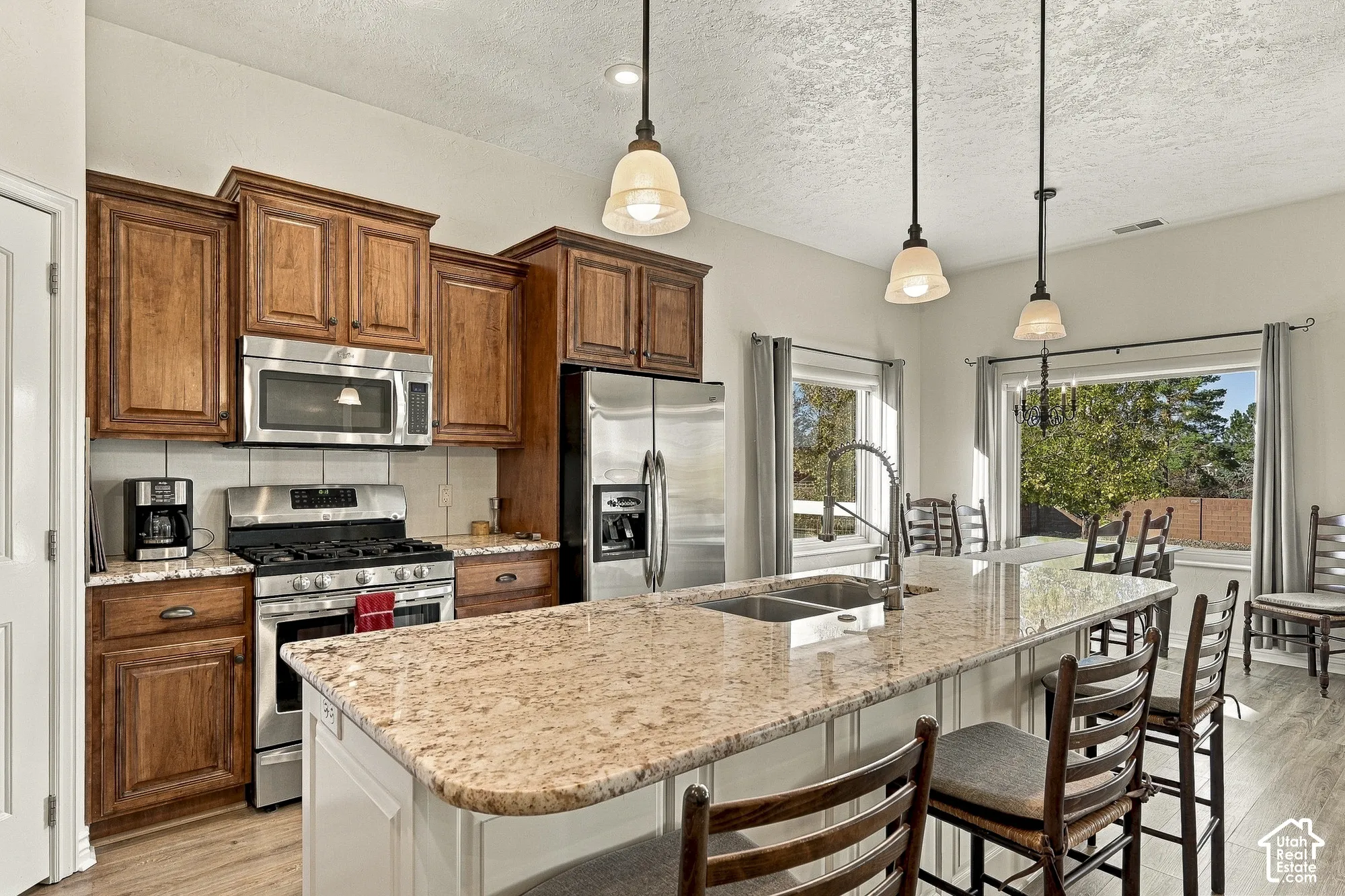 Kitchen featuring stained wood cabinetry, stone countertops, a central island with an integrated stainless steel dual-basin sink, stainless steel appliances, and three pendant lights