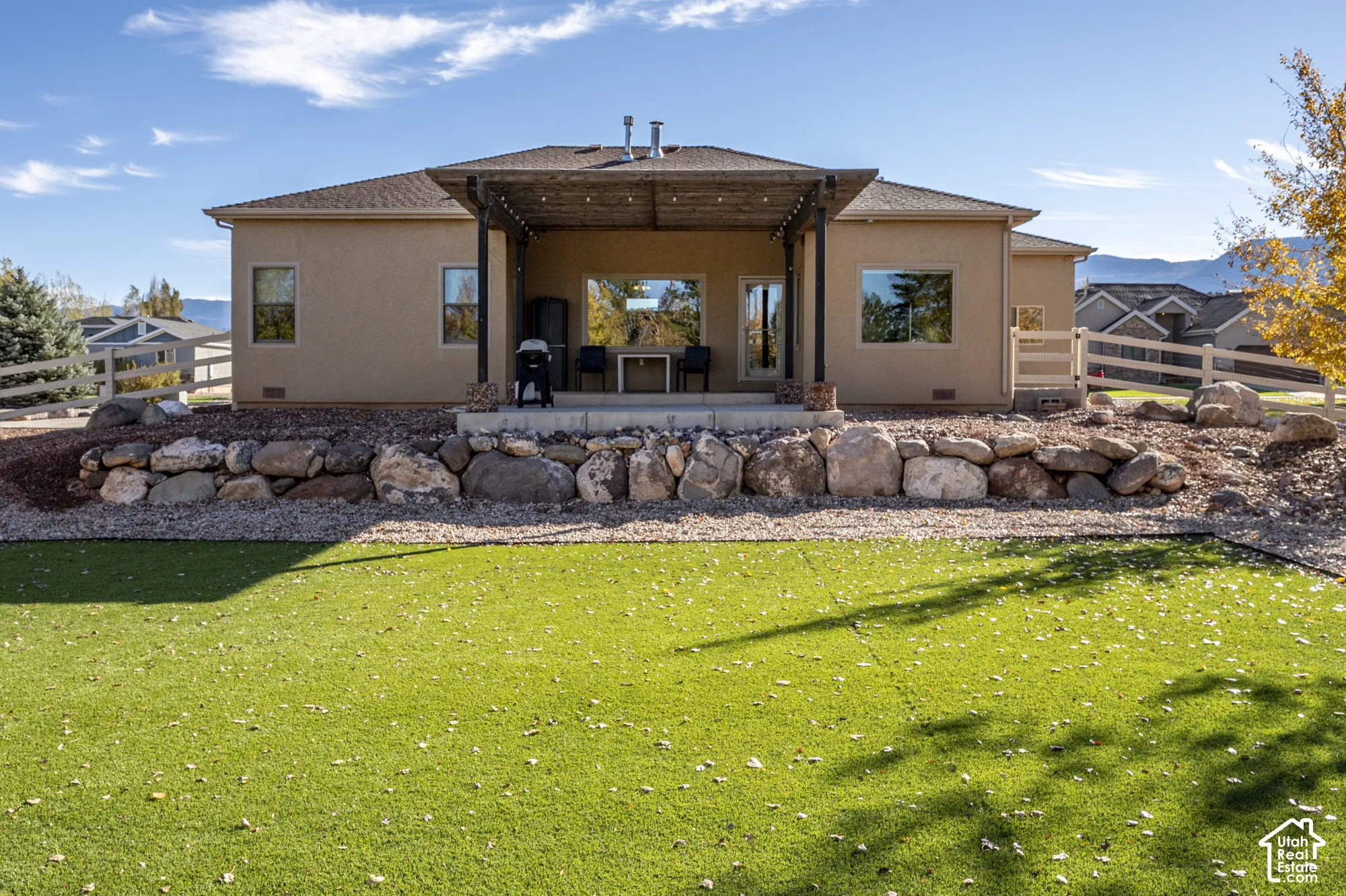 Stucco exterior featuring a covered patio with a wooden pergola, natural stone retaining wall, synthetic turf lawn, and white rail fencing
