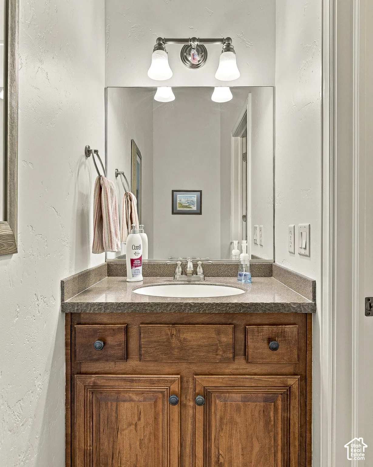 Vanity featuring a stone-composite countertop with an integrated oval sink, natural wood cabinetry, a large rectangular mirror, and dual-light wall-mounted fixture