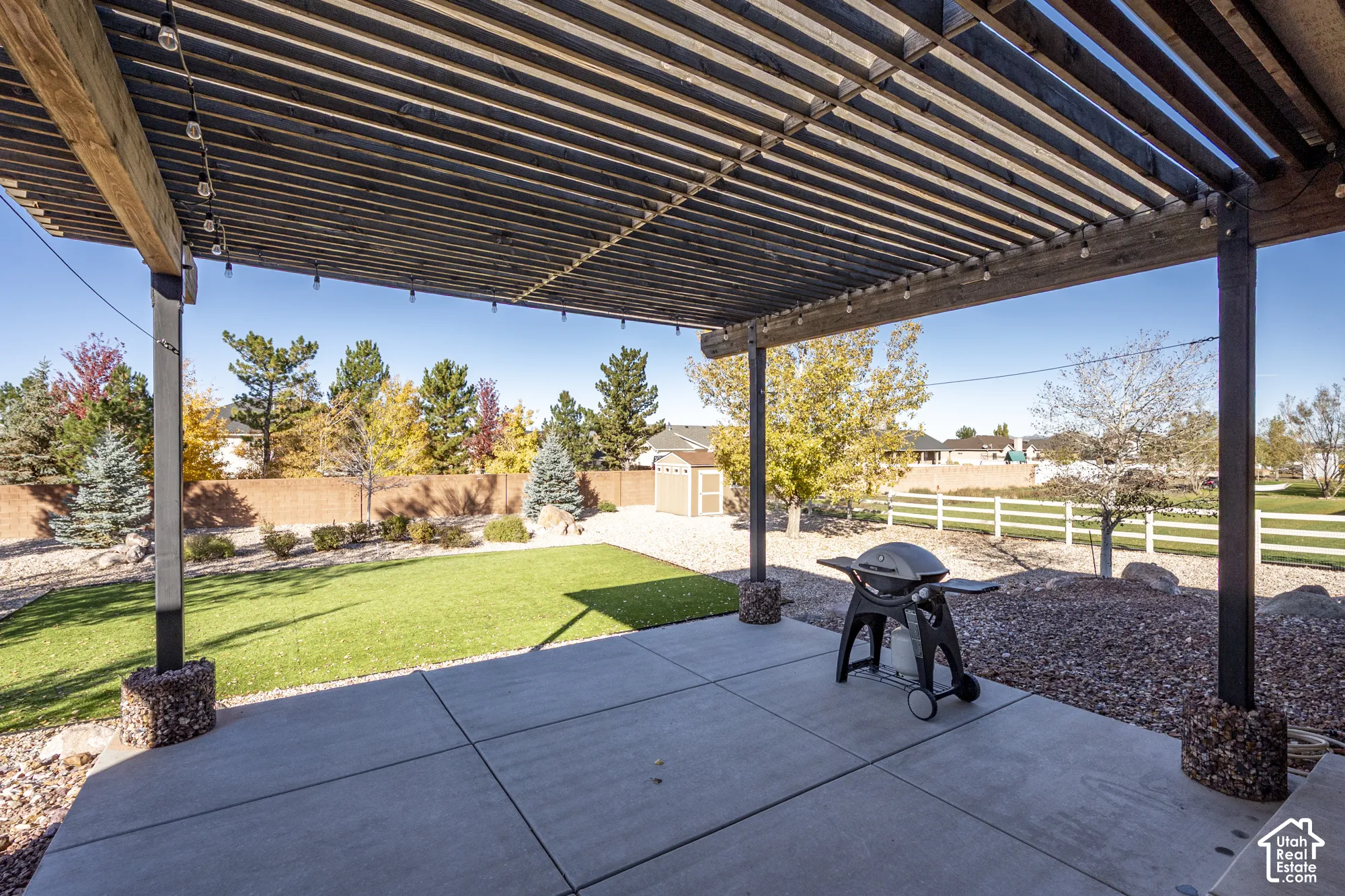Covered patio featuring a timber-slat pergola, concrete flooring, stone-base structural pillars, synthetic turf lawn, and decorative rock hardscaping