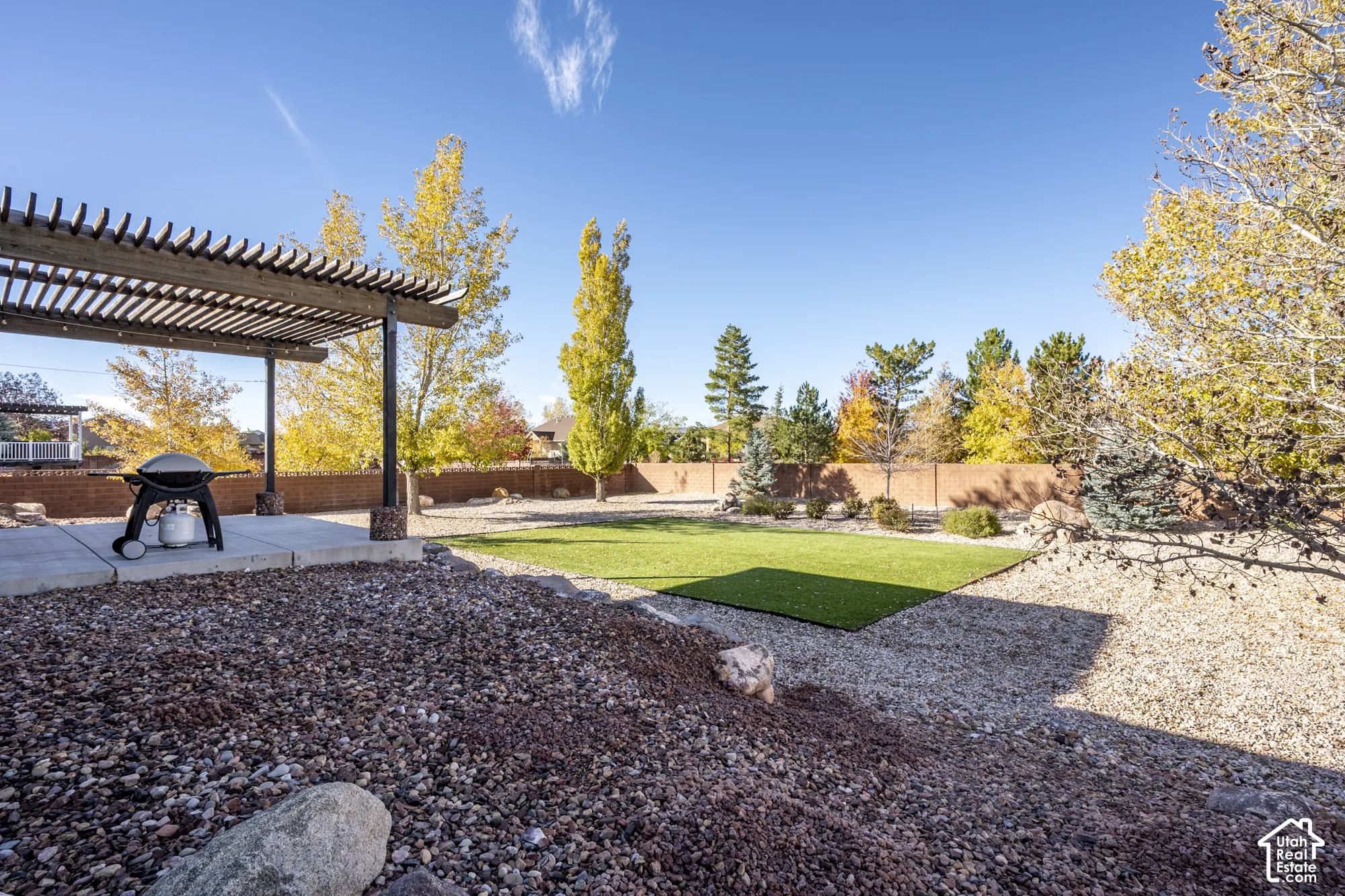 Backyard featuring a wood-slat pergola, concrete patio, synthetic turf lawn, decorative rock landscaping, and masonry perimeter wall