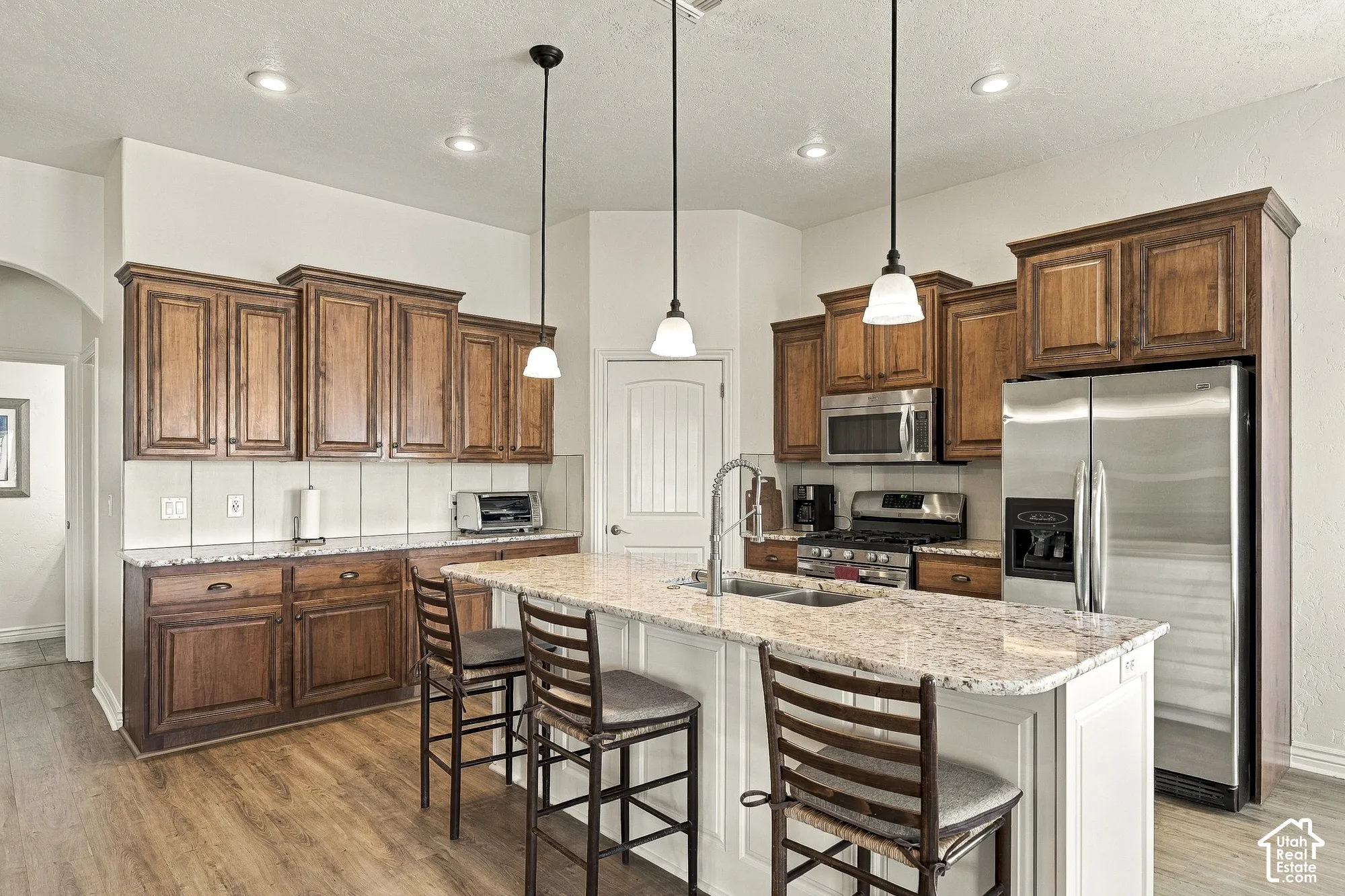 Kitchen featuring stained wood cabinetry, stone countertops, stainless steel appliances, a center island with undermount sink, and pendant lighting