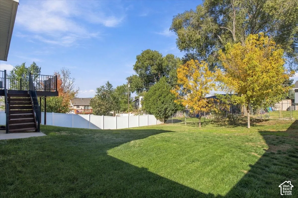 Expansive backyard featuring a manicured lawn, white vinyl fencing, mature trees, and an elevated deck with metal railings
