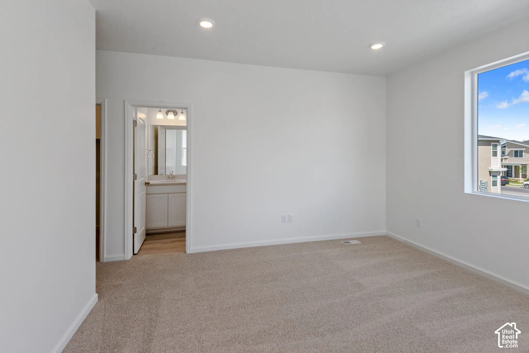 Neutral-toned wall-to-wall carpeting, recessed ceiling lighting, large window with natural light, and doorway revealing cabinetry with integrated sink